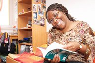 Eine Studentin sitzt auf einem Bett in einem Wohnheimzimmer und blickt lächelnd zum Betrachter. Sie hat ein aufgeschlagenes Buch in den Händen.