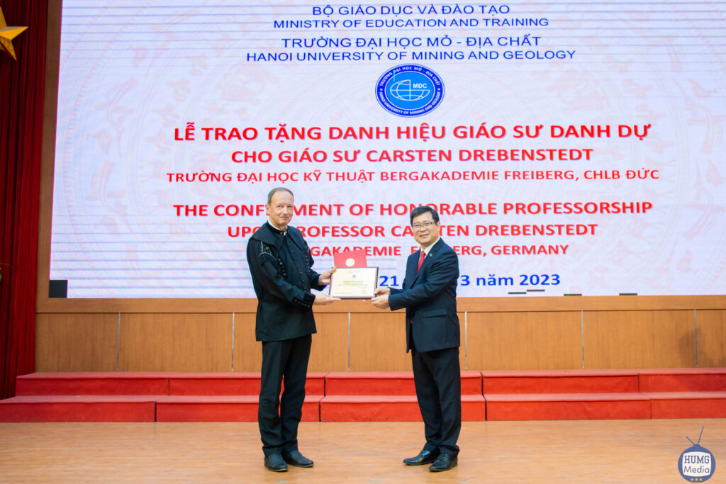 Zwei Professoren bei einer Ehrung. Der vietnamesische Professor übergibt dem deutschen Professor eine Urkunde, im Hintergrund ein großflächiger Text auf Vietnamesisch und Englisch